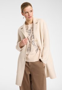 Donna che indossa un cappotto beige, un maglione ricamato con fiori e pantaloni marroni, che guarda di lato con una mano in tasca e l'altra sul bavero del cappotto.