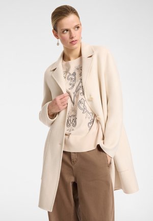 Donna che indossa un cappotto beige, un maglione ricamato con fiori e pantaloni marroni, che guarda di lato con una mano in tasca e l'altra sul bavero del cappotto.
