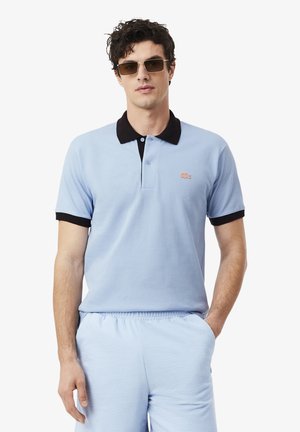 Hombre con camiseta polo azul claro con cuello y puños negros, pantalones cortos azul claro y gafas de sol rectangulares marrones, de pie con una mano en el bolsillo.