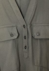 Graues Hemd mit Knopfleiste und V-Ausschnitt, zwei Brusttaschen und schwarzen Knöpfen. Es hat einen glatten, strukturierten Stoff mit dezenten Nähtendetails.