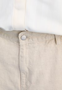Ljust beige linnebyxor med en texturerad yta, med en metallknapplåsning och en subtil sömdekoration längs midjan.