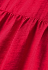 Roter Stoff mit einer zerknitterten Textur, der Sumpleiten aufweist. Glatte Oberfläche mit einem leichten Glanz, die hauptsächlich vertikale Linien zeigt.