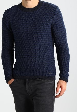 Pull en tricot texturé bleu marine avec un col rond et des poignets côtelés. Porté sur un jean foncé. Détail de marque discret près de l'ourlet.