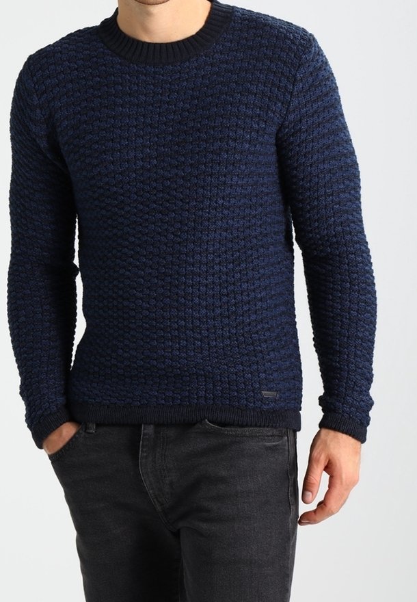 Dunkelblau gestrickter Pullover mit rundem Halsausschnitt und gerippten Bündchen. Über dunklen Jeans getragen. Dezentes Markenlogo nahe dem Saum.