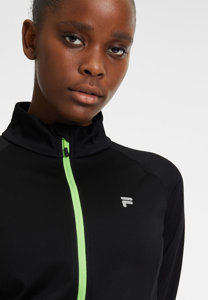 Giacca sportiva nera con colletto alto e zip verde. Presenta un logo argentato sul lato sinistro. Tessuto liscio, design aderente.