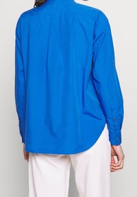 Langarmshirt aus lebhaftem blauen Baumwollstoff, mit abgerundetem Saum und geknöpften Manschetten. Glatte Textur mit taillierter Passform.