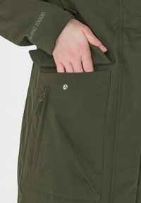 Olijfgroene jas met een hand in een zijzak. De stof heeft een waterafstotende afwerking met een gladdere textuur en zichtbare stiksels.