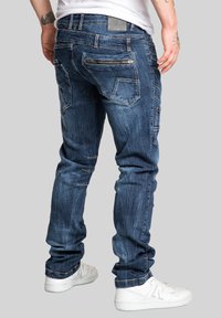 Dark blue denim jeans with a slim fit, detailed stitching, multiple pockets, and a zippered pocket on the back, paired with white sneakers.