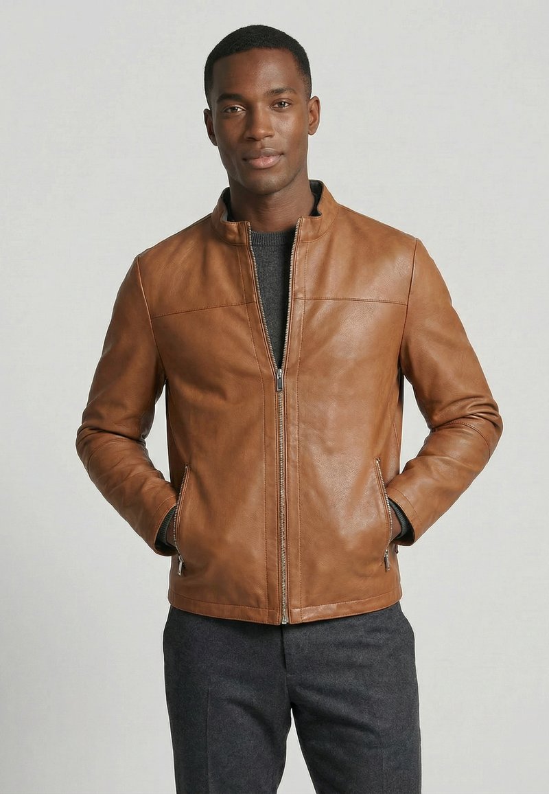 Mann steht mit den Händen in den Taschen, trägt eine braune Lederjacke mit Reißverschluss und dunkelgraue Hose vor neutralem Hintergrund.