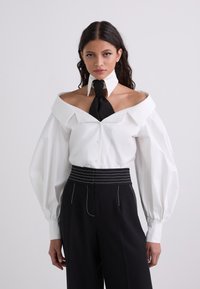 Camicia bianca off-the-shoulder con colletto a cravatta nera; presenta maniche voluminose e una chiusura frontale con bottoni, abbinata a pantaloni neri a vita alta.