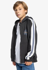Sudadera con cremallera negra y capucha con cordón, con franjas laterales en blanco y azul claro y un emblema en el pecho. Llevada sobre una camisa blanca.