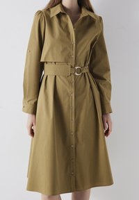 Mujer que lleva un vestido de camisa de manga larga en color caqui con botones y un cinturón ancho que presenta una hebilla de metal redonda en la cintura.