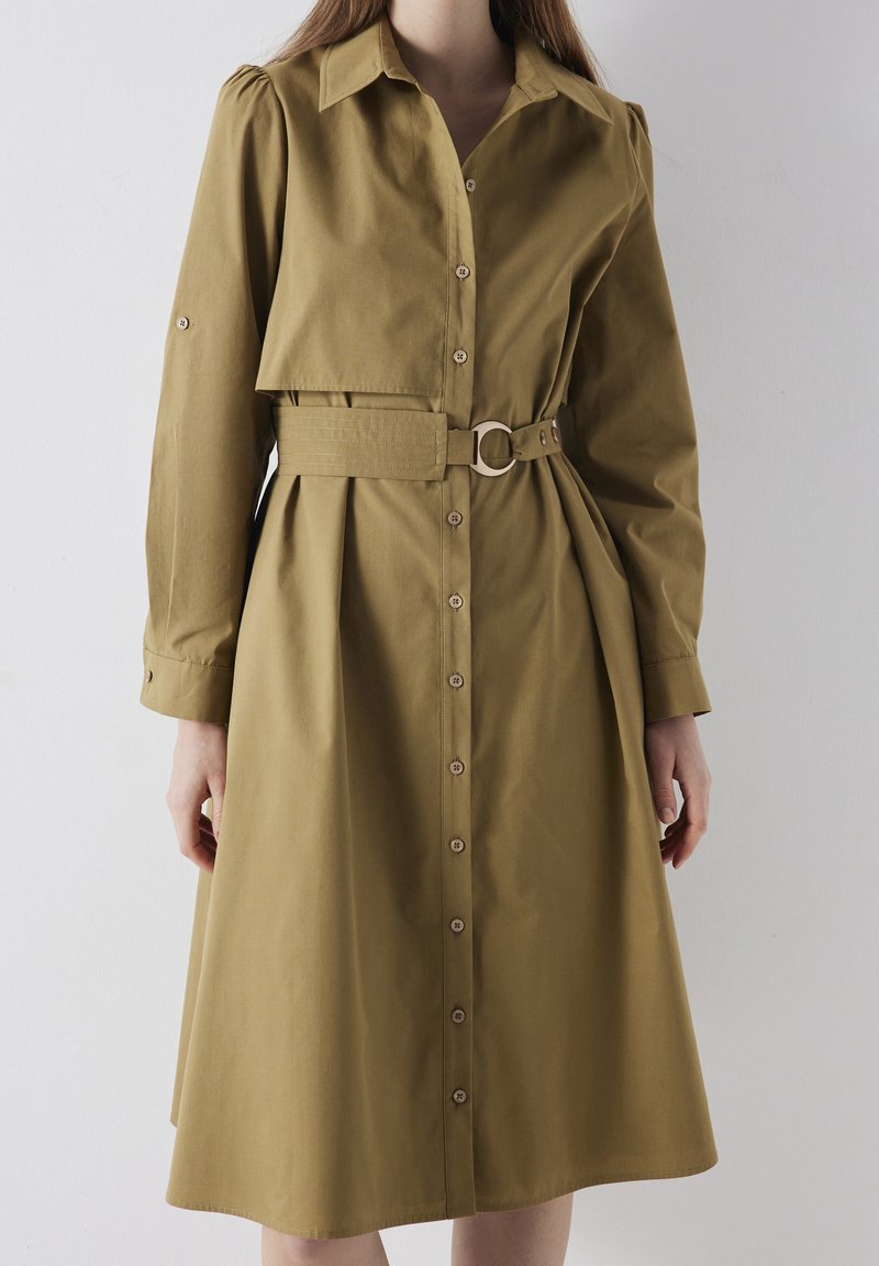 Mujer que lleva un vestido de camisa de manga larga en color caqui con botones y un cinturón ancho que presenta una hebilla de metal redonda en la cintura.