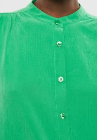 Close-up van een levendig groen shirt met een staande kraag en verschillende knopen, waarbij de textuur van de stof en de details van de couture zichtbaar zijn.