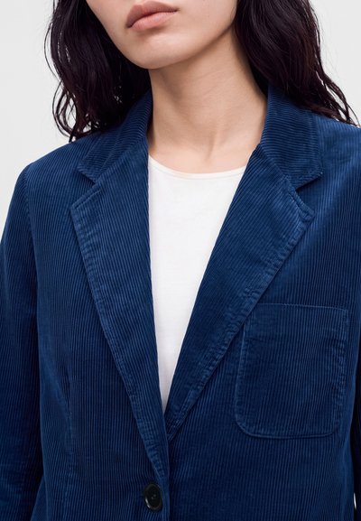 Femme portant un blazer bleu en velours côtelé sur une chemise blanche, montrant le col, le revers, la poche poitrine et les détails des boutons.