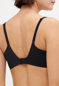 Frau trägt einen schwarzen BH mit verstellbaren Trägern und Hakenverschluss, von hinten vor einem einfarbigen Hintergrund gesehen.