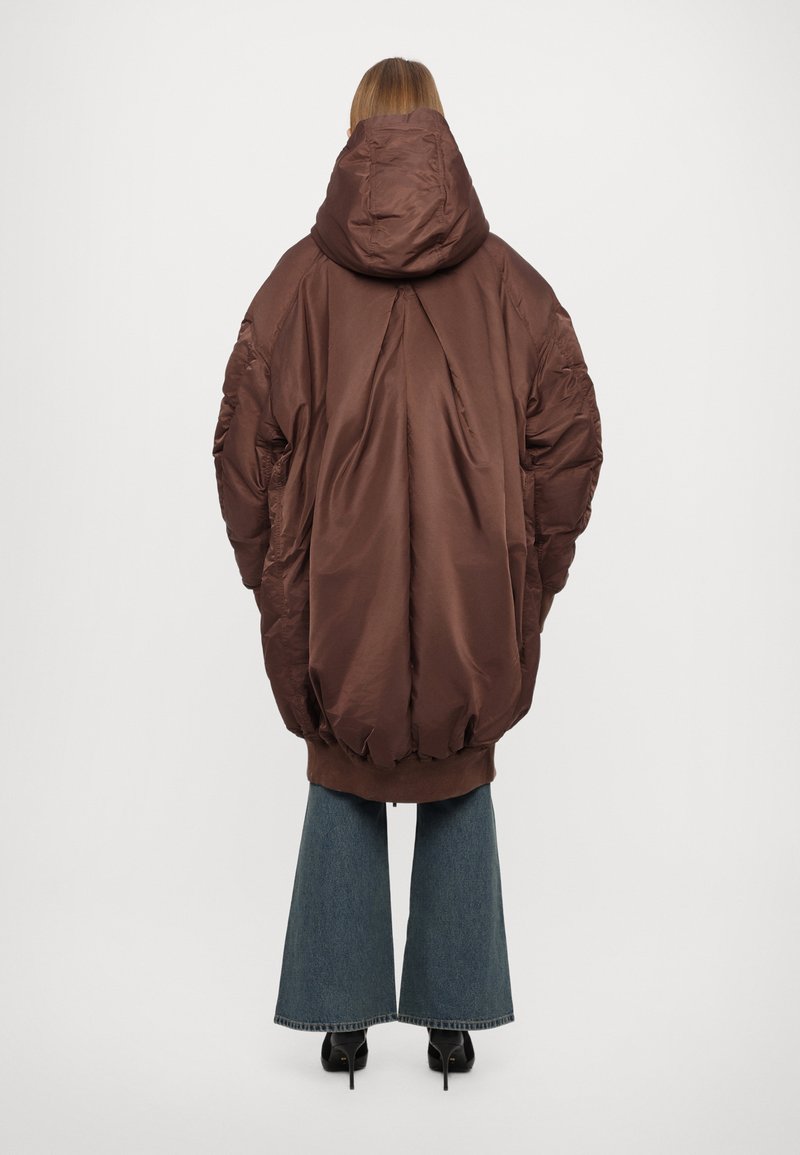 Übergroße braune Pufferjacke mit Kapuze, elastischen Bündchen und Saum, ausgestattet mit einer glatten Textur und einem Rückennähten-Detail. Kombiniert mit ausgestellten Jeans.