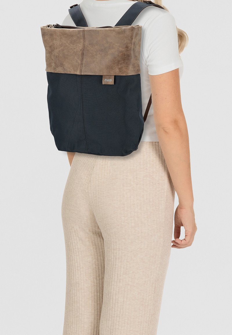 Zaino con parte superiore in pelle marrone e corpo in tessuto blu scuro, dotato di spallacci regolabili e chiusura con zip. Etichetta visibile sul lato.