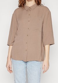 Donna che indossa una camicia button-up leggera e marrone chiaro con taschino e jeans azzurri, in piedi contro uno sfondo neutro.