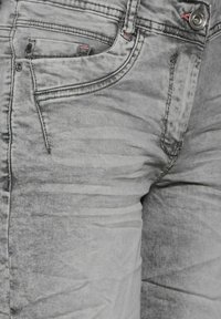 Jeans en denim gris avec un lavage délavé, des coutures contrastantes et des poches avant avec des accents en métal. Le tissu a une apparence douce et texturée.