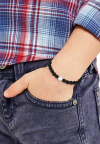 Bracciale di perline nere con texture opaca liscia e un'unica perla argentata lucida come accentuazione, indossato su una mano poggiata in una tasca di jeans scuri.