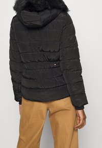 Černá péřová bunda s kapucí lemovanou kožešinou, prošívaným designem, elastickým pasem a kapsami na zip, nošená s béžovými kalhotami.