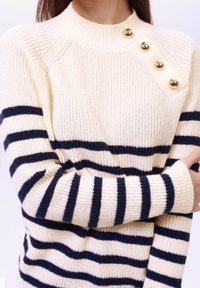 Pull en tricot crème avec des rayures navy, col montant et trois boutons dorés à l'épaule. Texture côtelée et manches longues.