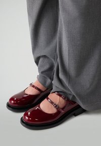 Schimmernde bordeauxrote Mary-Jane-Schuhe mit doppeltem Riemen, getragen mit lockeren grauen Hosen, stehend auf einem hellgrauen Boden.