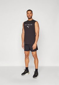 Črna majica brez rokavov z logotipom "HYROX", kombinirana s črnimi kratkimi hlačami in črnimi športnimi čevlji z zelenimi poudarki, stoji na svetlem ozadju.