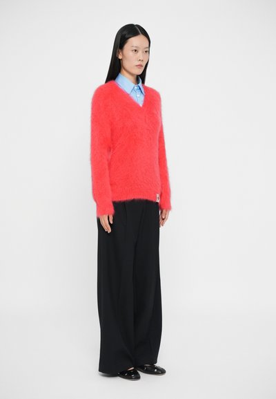 Maglione girocollo morbido di un vivace rosa, realizzato in materiale soffice. Indossato sopra una camicia azzurra con colletto, abbinato a pantaloni neri larghi.