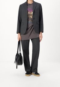 Grå randig kavaj över en grafisk t-shirt, ihop med matchande byxor och svarta sneakers. Håller en svart, dragsko-handväska.