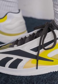 Vita sneakers med svarta och gula accenter, med en texturerad överdel, tätt snörda och en solid gummisula med en strömlinjeformad design.