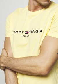 T-shirt en coton jaune avec le logo "TOMMY HILFIGER" en noir et un accent rectangulaire coloré, col rond et manches courtes.