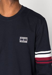 Navy Langarm-T-Shirt aus Baumwolle mit einem kleinen gestickten Logo auf der Brust und gestreiften Akzenten in Rot, Weiß und Blau an den Ärmeln.