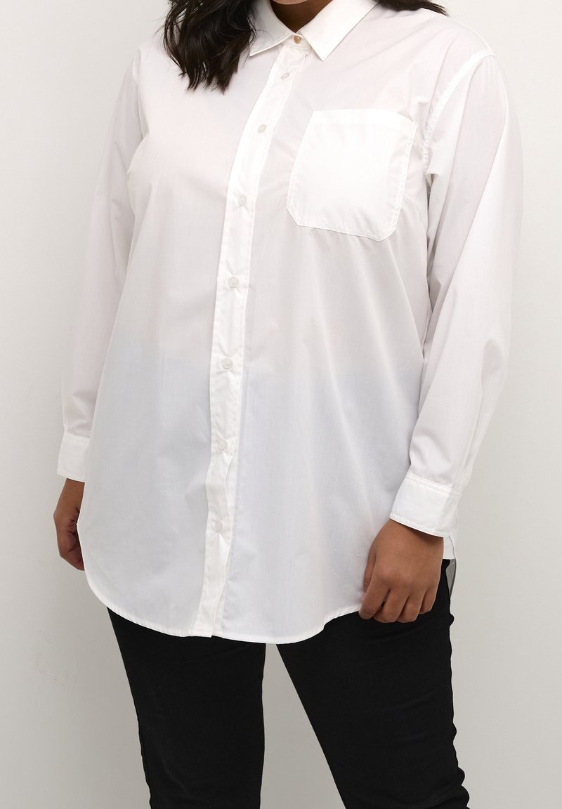Femme portant une chemise blanche à manches longues avec une poche poitrine et un pantalon noir, debout devant un fond uni.