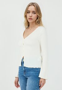 Cardigan bianco a coste con maniche lunghe, scollatura a V profonda e orlo scallopato, caratterizzato da una chiusura con bottoni e una consistenza morbida. Indossato con jeans blu.