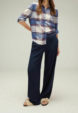 Vrouw draagt een blauw-wit geruit overhemd met opgerolde mouwen, losse donkerblauwe broek en beige zachte pantoffels, staand met handen in de zakken.