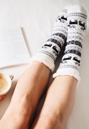Calze invernali lavorate a maglia bianche e nere, con un motivo a cervo e polsini strutturati. Pelle delle gambe liscia e una tazza con una bevanda nelle vicinanze.