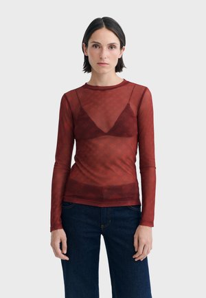 Femme aux cheveux courts et foncés portant un haut rouge transparent à manches longues par-dessus un soutien-gorge assorti et un jean bleu foncé, debout devant un fond uni.