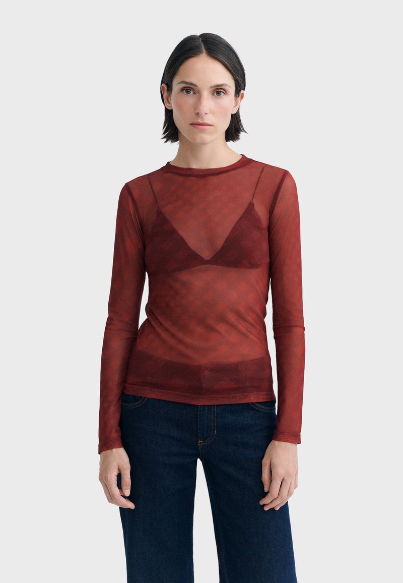 Femme aux cheveux courts et foncés portant un haut rouge transparent à manches longues par-dessus un soutien-gorge assorti et un jean bleu foncé, debout devant un fond uni.