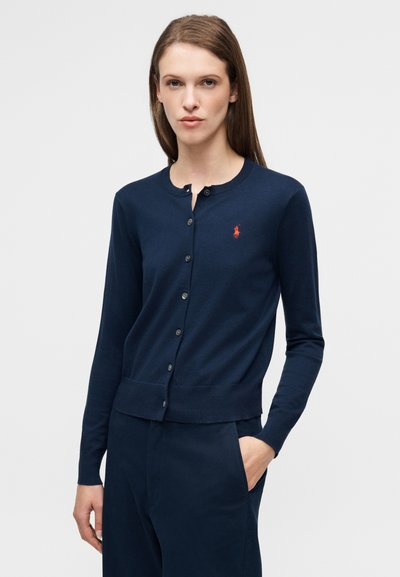 Κάρντιγκαν σε σκούρο μπλε ναυτικό χρώμα από λείο ύφασμα, με στρογγυλή λαιμόκοψη, κουμπιά μπροστά και μικρό κόκκινο λογότυπο στο αριστερό μέρος του στήθους.