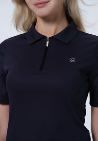 Navyblaues Poloshirt aus glattem Material, mit einem halben Reißverschluss vorne und einem kleinen silbernen Logo auf der linken Brust.