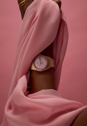 Montre rose avec des accents dorés et un cadran rose, comportant un affichage de la date et un bracelet en silicone, drapée dans un fond de tissu rose fluide.
