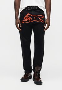 Μαύρο τζιν παντελόνι με μεγάλο πορτοκαλί γραφικό μιας γάτας στον αριστερό μηρό. Διαθέτει ίσια γραμμή και πέντε παραδοσιακές τσέπες.