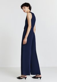 Combinaison bleu marine avec un buste ajusté et des jambes larges, présentant une découpe dans le dos et une texture de tissu. Accompagnée de sandales à talons noires.