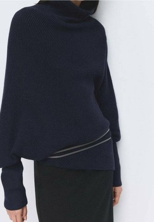 Femme portant un pull à col roulé côtelé bleu marine foncé à manches longues et une jupe noire, debout contre un mur blanc.