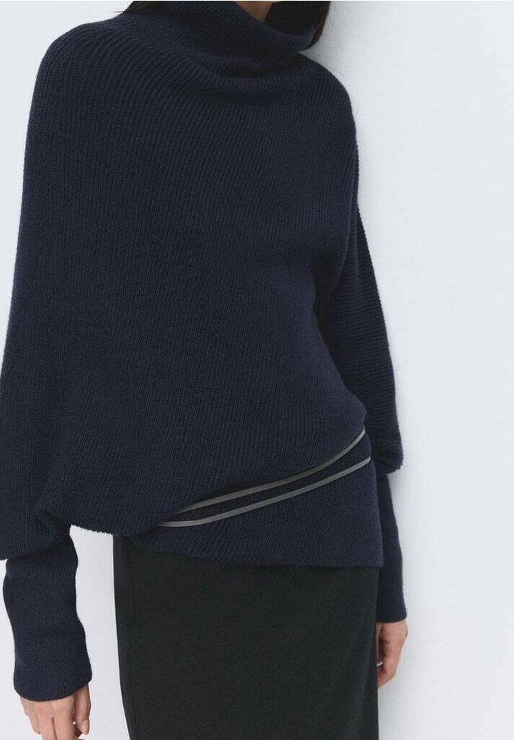 Femme portant un pull à col roulé côtelé bleu marine foncé à manches longues et une jupe noire, debout contre un mur blanc.
