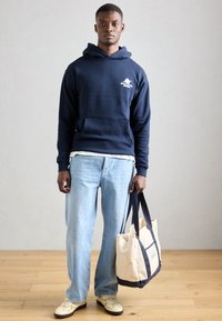 Hanorac navy cu buzunar tip kangaroo, blugi deschiși, și geantă crem cu accente navy. Modelul poartă adidași ușori.