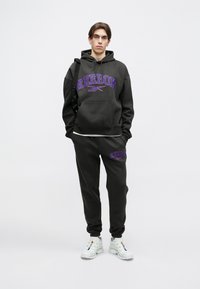 Tumši pelēks Reebok džemperis un sporta bikses ar lielu purpursarkanu logotipu priekšpusē. Apģērbs ir brīvs un to papildina baltas kedas.