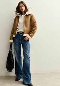 Giacca in finta shearling marrone con fodera bianca, maglione bianco sotto, jeans a zampa d'elefante blu e borsa nera. Tessuto liscio e design casual.